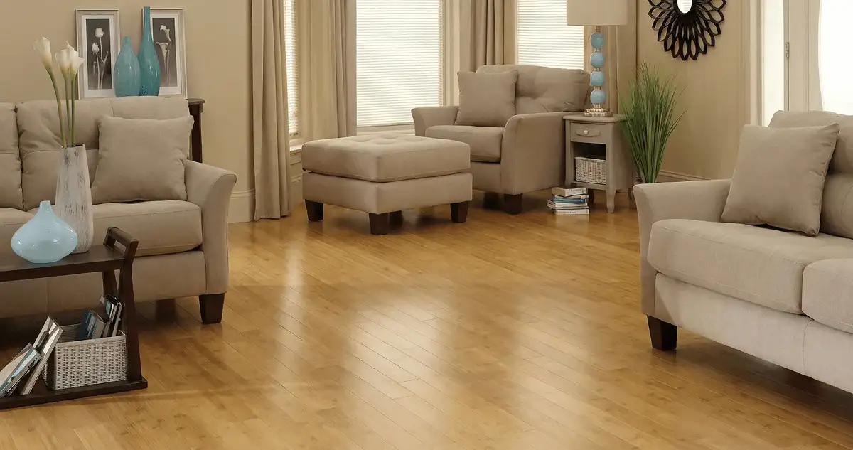A living room with professionally installed bamboo flooring, beige sofas, a matching ottoman, a side table with a blue lamp, vases, framed pictures, and large windows with blinds and curtains in Des Moines, IA