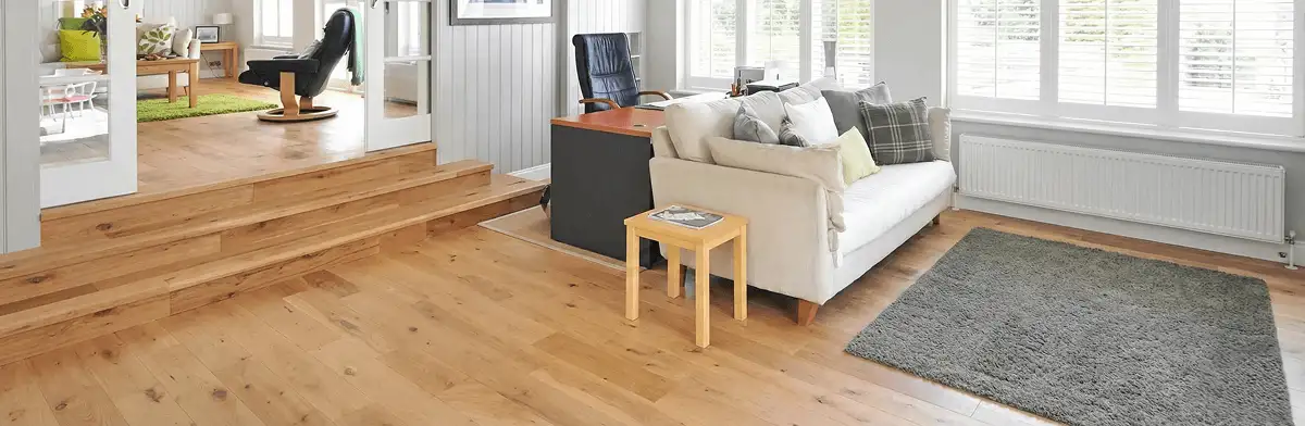 A modern living room with professionally installed laminate flooring, a beige sofa, a small side table, a gray rug, and large windows with white shutters in Des Moines, IA